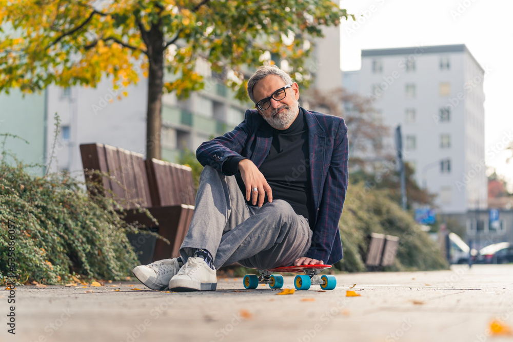 Proud and confident caucasian gray-haired man in his 60s dressed in business casual way sitting cross-legged on modern neon orange skateboard and looking at camera. Sport for everyone. High quality Proud and confident caucasian gray-haired man in his 60s dressed in business casual way sitting cross-legged on modern neon orange skateboard and looking at camera. Sport for everyone. High quality