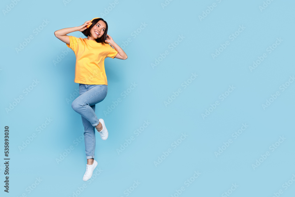 Full length photo of young korean girl jump air trampoline listen headphones music look interested empty space isolated on blue color background