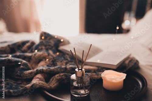 Bamboo sticks in bottle with scented candles and open book on wooden tray with knitted sweater fabric cloth in bed closeup. Home aroma.