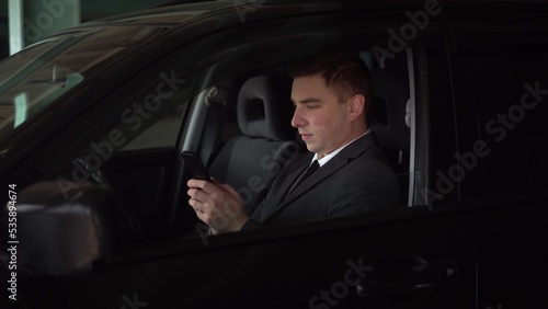 Young businessman chatting on the phone while sitting in the car in the parking lot. A man in a suit with a phone in his hand in the front seat. © Vital9c