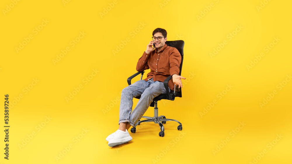 Male Talking On Phone Sitting In Office Chair, Yellow Background