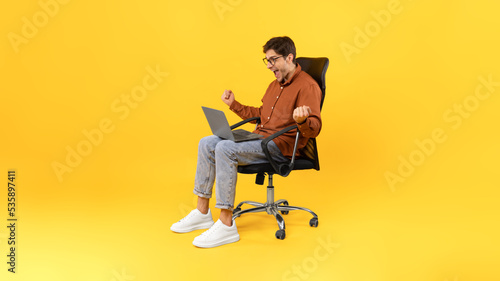Joyful Guy Shaking Fists Using Laptop Sitting Over Yellow Background