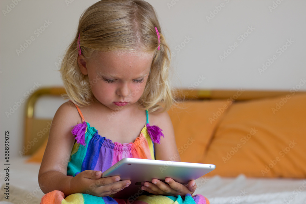 Little blond girl sitting on bed and holding tablet,toddler using internet with gadget,kid addicted to tablet and looking cartoons,3 year old child studiyng online with developmental games. Little blond girl sitting on bed and holding tablet,toddler using internet with gadget,kid addicted to tablet and looking cartoons,3 year old child studiyng online with developmental games.
