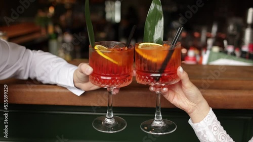 hands of woman and man are clinking, cheers with glasses of Spritz cocktail. Couple celebrating wedding, anniversary with Aperol spritz cocktails, with orange and greens Refreshing alcoholic drink.