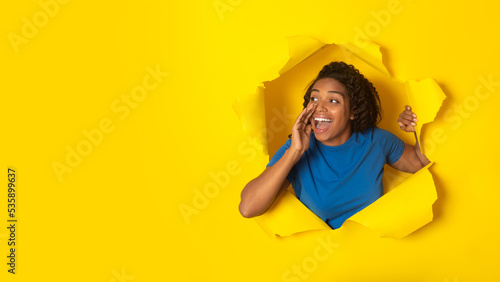 Black Female Shouting Through Hole In Torn Yellow Paper Background photo