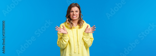 Young woman describing disgusting story, grimacing from aversion and dislike, raising hands and squinting bothered, feel disgust from awful smell or cringy thing she see, blue background photo