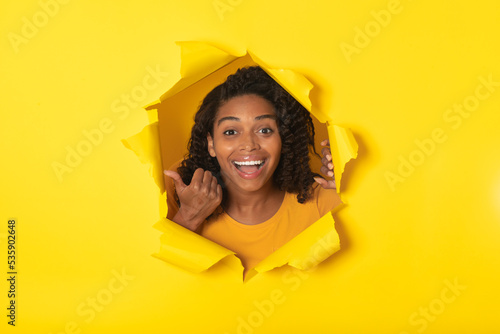 Excited Black Female Looking Through Hole In Paper, Yellow Background