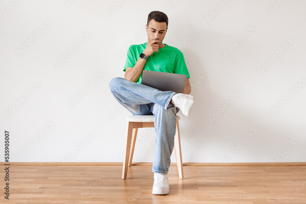 Pensive arab man using laptop and thinking, browsing internet while sitting on chair against white wall at home Pensive arab man using laptop and thinking, browsing internet while sitting on chair against white wall at home