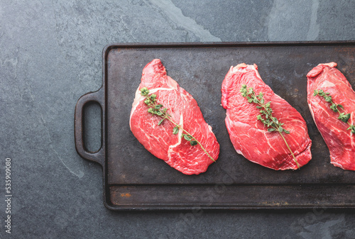 Raw beef steaks with herbs and spices on cast iron frying board, top view