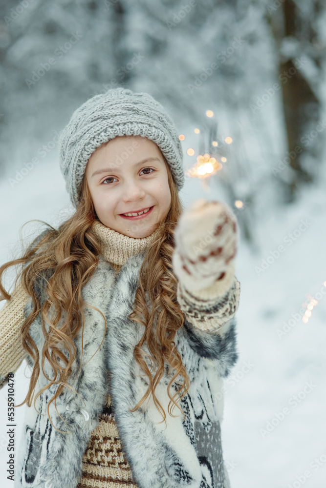 Young happy smiling girl outdoors in white cold snow covered winter forest or park with sparklers, dressed in knitted hat, sweater, fur vest and mittens - seasonal portrait with festive christmas mood Young happy smiling girl outdoors in white cold snow covered winter forest or park with sparklers, dressed in knitted hat, sweater, fur vest and mittens - seasonal portrait with festive christmas mood
