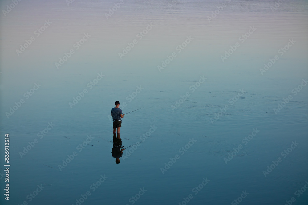Fototapeta premium A fisherman stands in the water and catches fish with a fishing rod. River or lake, no waves