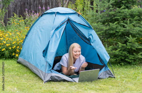 A young girl lying on the grass in the tourist tent with a laptop and communicates through a video call.