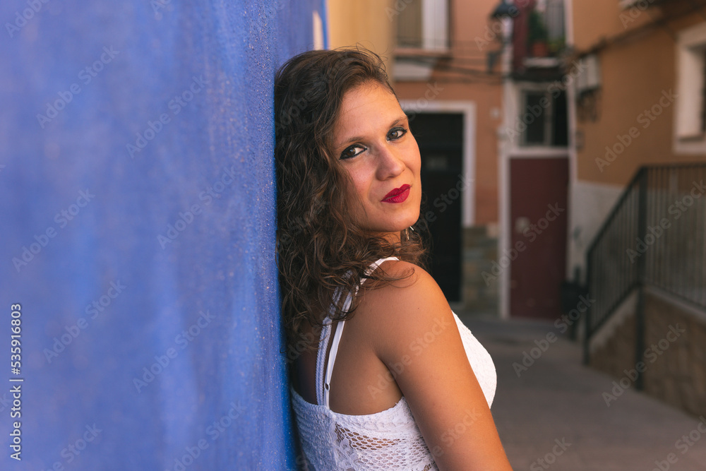 Serious brunette woman looking at the camera and leaning her back against a blue wall in the narrow and colorful streets of Villajoyosa, Alicante, Spain.