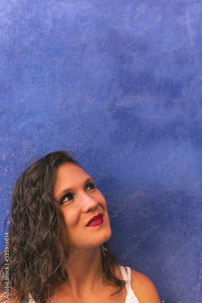 Image vertical of a brunette woman looking up while grimacing with a smile leaning against a blue wall inside the narrow colorful streets of Villajoyosa, Alicante, Spain.w Image vertical of a brunette woman looking up while grimacing with a smile leaning against a blue wall inside the narrow colorful streets of Villajoyosa, Alicante, Spain.w