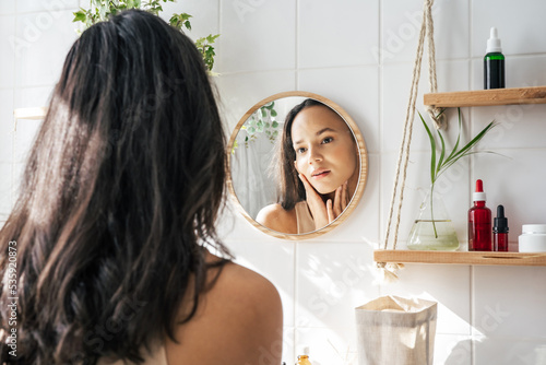 Beautiful woman with dark skin looking in the mirror in white eco friendly bathroom. Wooden shleves and reusable cosmetics bottles. Wellnes concept