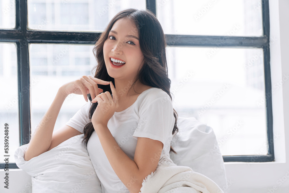 Lovely asian woman casual attire sitting with pillow beside windows relax peaceful in morning at home. Cheerful healthy young girl resting on white pillow with sunlight looking something in bed room. Lovely asian woman casual attire sitting with pillow beside windows relax peaceful in morning at home. Cheerful healthy young girl resting on white pillow with sunlight looking something in bed room.