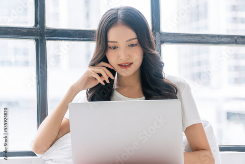 Happy asian casual woman reading news writing online with laptop or shopping comfortable at home. Freelance young girl using notebook relax, work sitting with pillow beside window.Lifestyle Technology