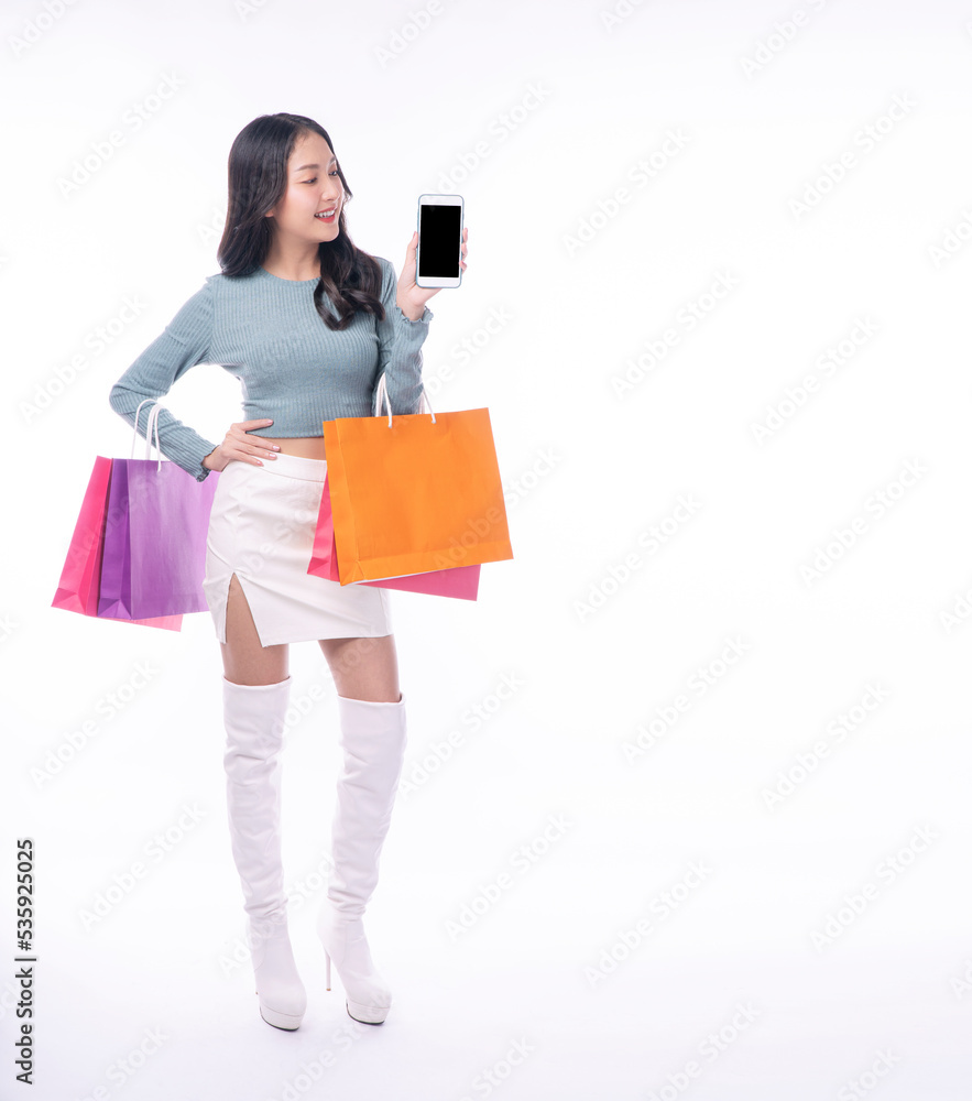 Cheerful excited asian woman showing mobile phone screen carrying shopping bags full body standing on white background. Joyful model young girl surprise application shopping online over isolated. Cheerful excited asian woman showing mobile phone screen carrying shopping bags full body standing on white background. Joyful model young girl surprise application shopping online over isolated.
