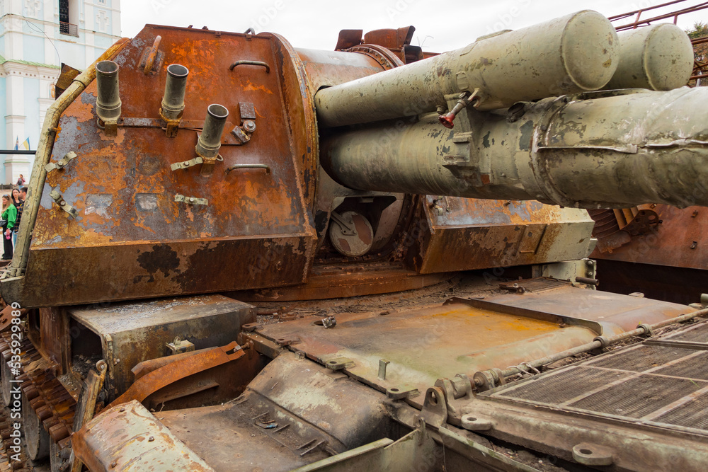 A destroyed rusty military tank stands outdoors on the street. Battle. Damage. Fight. Heavy. Defense. Armored. Shelling. Death. Explosion. Victim. Rust. Wrecked. Artillery. Outdoors. Remains