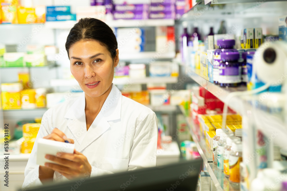 Oriental female pharmacist standing at counter in chemist shop and writing recipe.