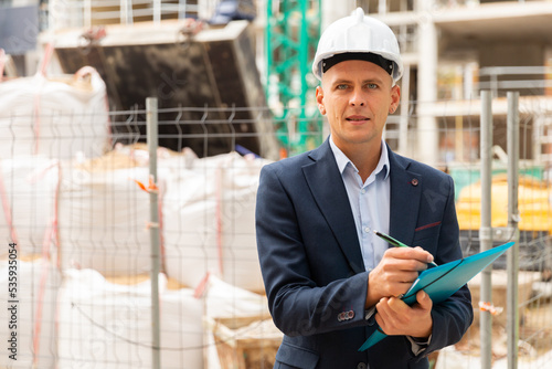 Engineer in suit and hardhat standing in construction area with paper folder in hands.