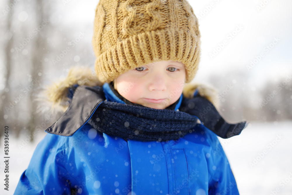 Portrait of little upset boy during walk in snowy winter park. Kid dressed in warm clothes, hat and scarf. Active winter outdoors leisure for child Portrait of little upset boy during walk in snowy winter park. Kid dressed in warm clothes, hat and scarf. Active winter outdoors leisure for child