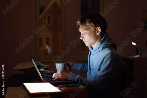 Concentrated teenager studying late night at home office with laptop and tablet.