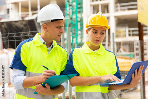 Man and woman builders in uniform and hardhats discussing while standing in construction site. Woman using laptop, man holding folder with documentation.