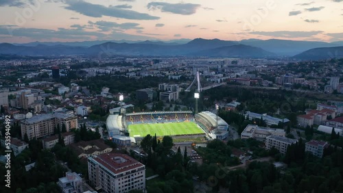 Cityscape featuring a stadium with green grass soccer field (football pitch) illuminated by floodlights at night, in the dark city of Podgorica, Montenegro, under colorful sunset sky in the evening.