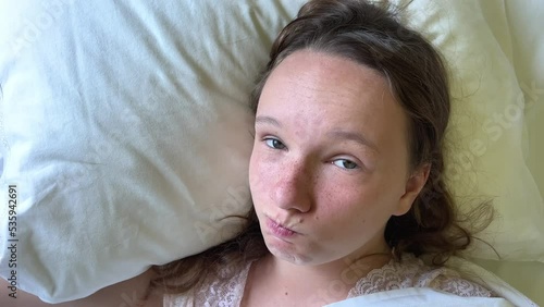 a teenage girl lies on a white pillow and smiles looks into the frame she has run out of either a cold or an allergy she is happy recovered shows class with her eyes. High quality 4k footage