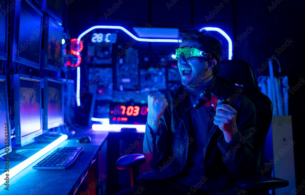 Gamer man with hitech clothes and eye glasses show action of excited and happy during play computer game in room with dark and multi-color light. Gamer man with hitech clothes and eye glasses show action of excited and happy during play computer game in room with dark and multi-color light.