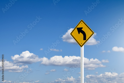 Traffic sign; left arrow curve sign on white pole by the main road with blue sky background, soft and selective focus.