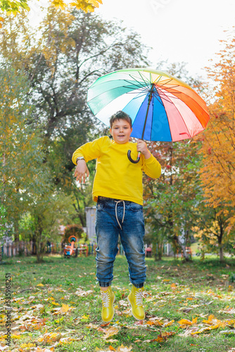 Full length portrait of a cute boy dressed in yellow long sleeve and yellow rubber boots jumping with a colorful umbrella. A child in the autumn park posing with rainbow umbrella.