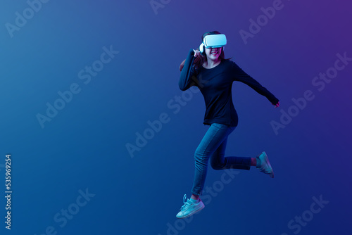 Young Asian woman wearing VR headset with experience playing video game and jumping levitating in the air on futuristic purple cyberpunk neon light background. Metaverse technology concept.