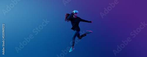 Young Asian woman wearing VR headset with experience playing video game and jumping levitating in the air on futuristic purple cyberpunk neon light banner background. Metaverse technology concept.