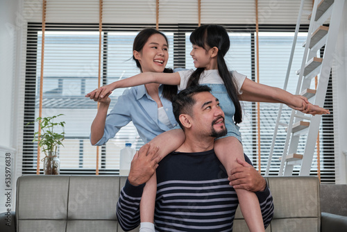 Asian Thai family together, dad plays and teases with daughter and mum by carrying and holding girl on shoulders in home living room, happy leisure times, lovely weekend, wellbeing domestic lifestyle.