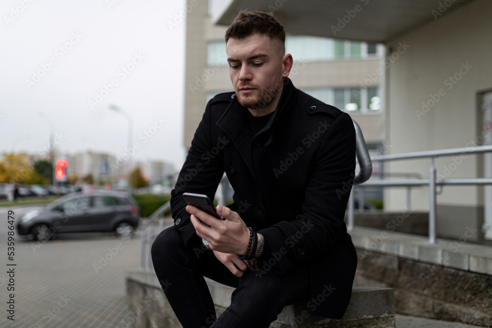 brutal young man in a black coat with a mobile phone on the background of the office center brutal young man in a black coat with a mobile phone on the background of the office center