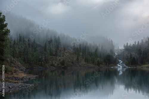 Fototapeta Naklejka Na Ścianę i Meble -  Mountain creek flows from forest hills into glacial lake in mysterious fog. Small river and coniferous trees reflected in calm alpine lake in early morning. Tranquil misty scenery with mountain lake.