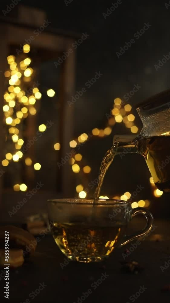 in the dark, by the light of garlands, hot tea is poured into a transparent mug from a glass teapot in the dark, by the light of garlands, hot tea is poured into a transparent mug from a glass teapot