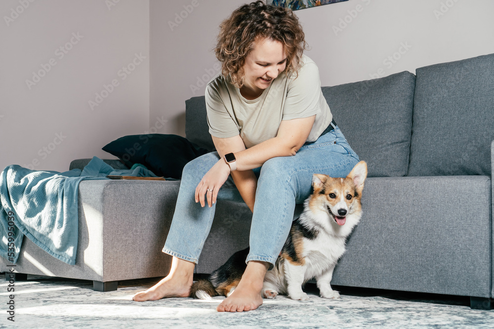 Curly haired glad, cheerful barefoot woman with dog corgi sitting, relaxing and playing on floor in comfortable living room, near sofa. Active and healthy animal, healthcare. Weekend leisure activity