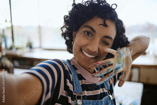 Woman artist taking selfie, painter in her art workshop and happy with a hand full of paint. Young, Indian professional creative smile in self portrait, having fun painting and working.