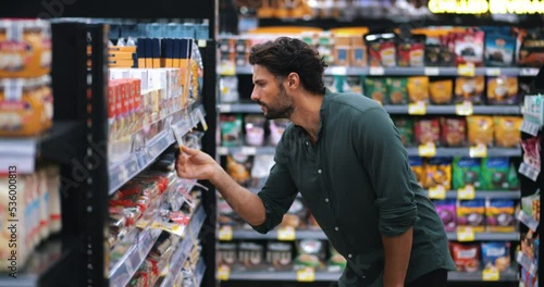Thoughtful bearded attractive young man is shopping in the grocery department of a supermarket. Calculating expensive prices, looking aside on shelf with food, standing with a trolley in a supermarket