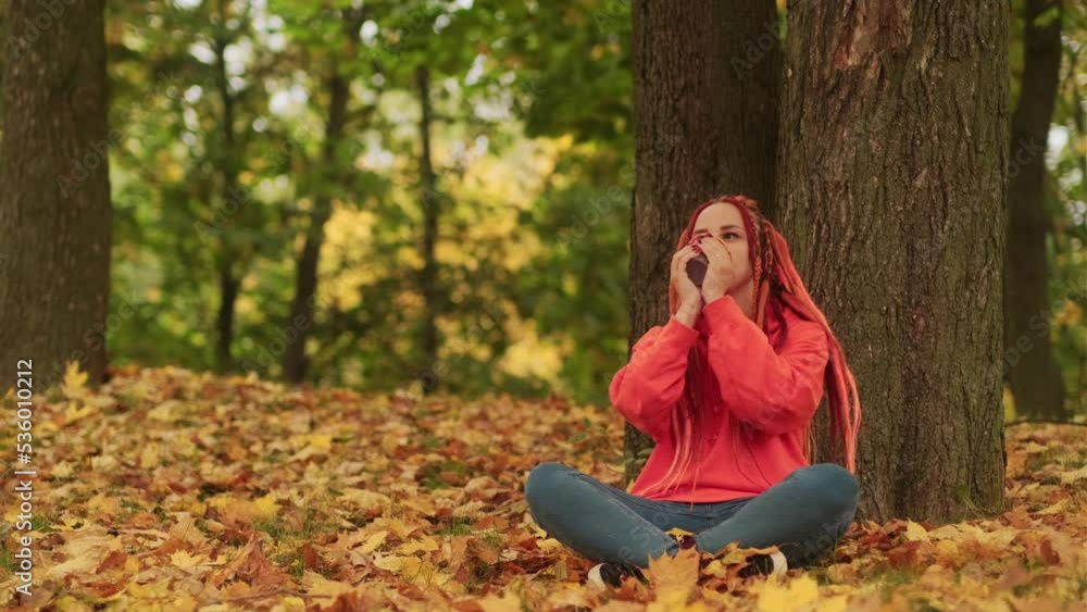 Young woman with dreadlocks drinking coffee from cup, sitting near tree in golden autumn. Vivid female with colourful hairstyle enjoying hot drink in nature. Young woman with dreadlocks drinking coffee from cup, sitting near tree in golden autumn. Vivid female with colourful hairstyle enjoying hot drink in nature.