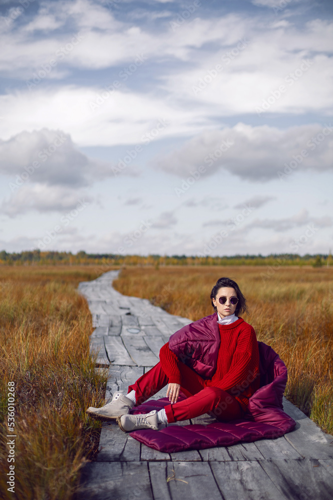 portrait of a fashionable beautiful brunette woman sit on an ecological path in the marshes in autumn in a red sweater, hat and an unusual scarf. in Yelnya in Belarus.