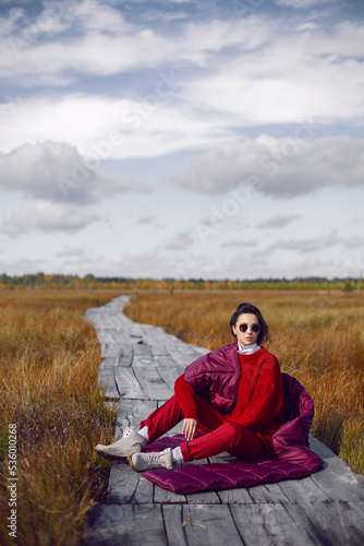 portrait of a fashionable beautiful brunette woman sit on an ecological path in the marshes in autumn in a red sweater, hat and an unusual scarf. in Yelnya in Belarus. © saulich84