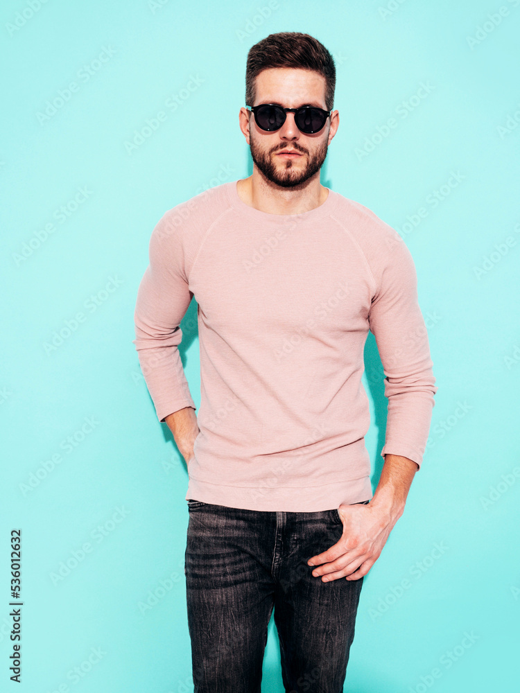 Fototapeta premium Portrait of handsome model. Sexy stylish man dressed in pink sweater and jeans. Fashion hipster male posing near blue wall in studio. In sunglasses. Isolated