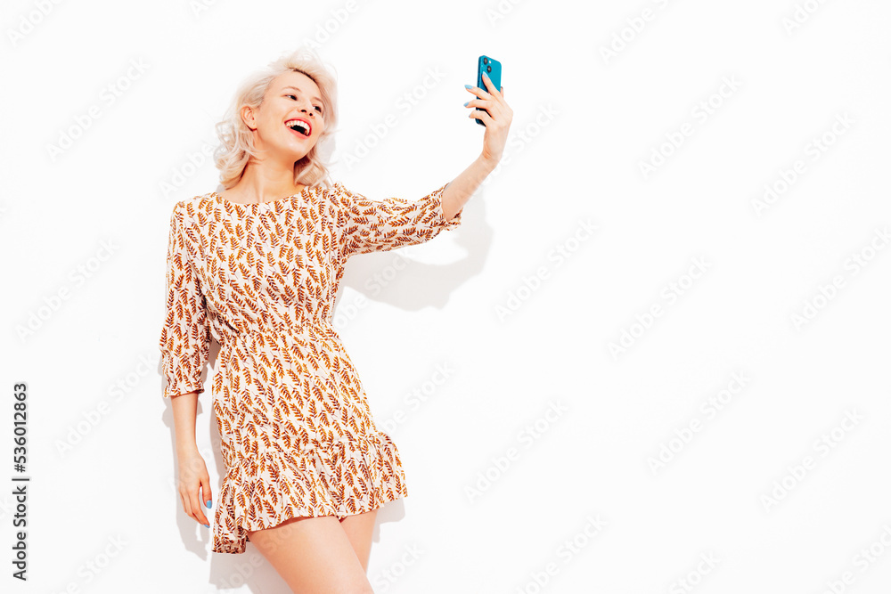 Portrait of young beautiful smiling female in trendy summer dress. Sexy carefree blond woman posing near white wall in studio. Positive model having fun indoors. Cheerful and happy. Taking selfie