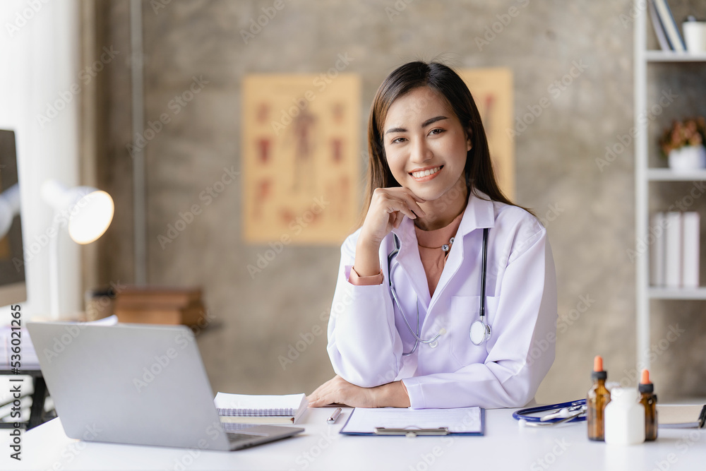 Asian female doctor sitting at a hospital desk, giving health care and disease prevention advice, convenient online services to patients and smiling, writing prescriptions to order medicine.