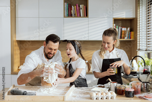 Adorable family in modern kitchen coking baking together. Happy father pouring flour delighted daughter holding sieve talking watching process. Happy mother looking at dad and girl opening jam.
