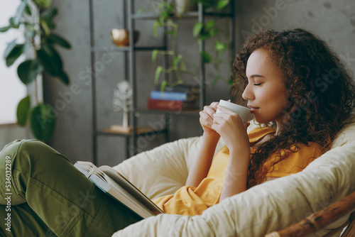 Sideways young woman of African American ethnicity wear t-shirt drink coffee read book close eyes sits in armchair stay at home flat rest relax spend free spare time in living room indoor grey wall. photo